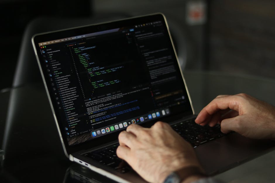 A close-up of a person's hands working on a laptop displaying lines of code, with a dark background. The laptop is placed on a reflective surface, and the screen shows a coding interface with various